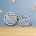 Two tins of diaper cream on a wooden surface with a blue background and gold stars.